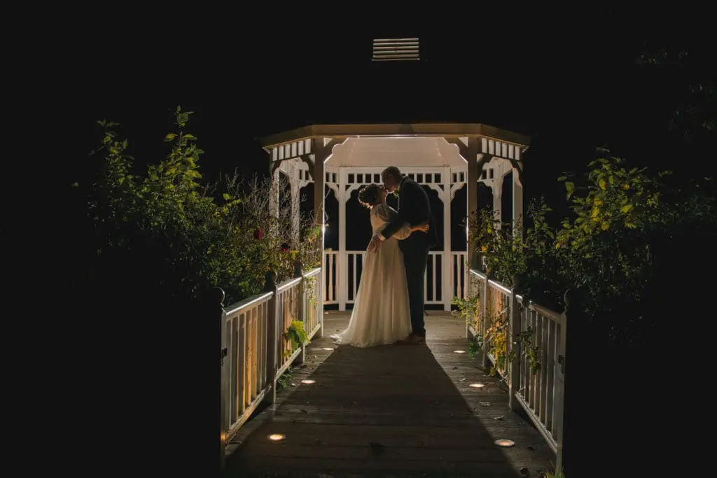 A bridal couple kiss in the distance in an arbour, it is night time and the scene is beautifully lit