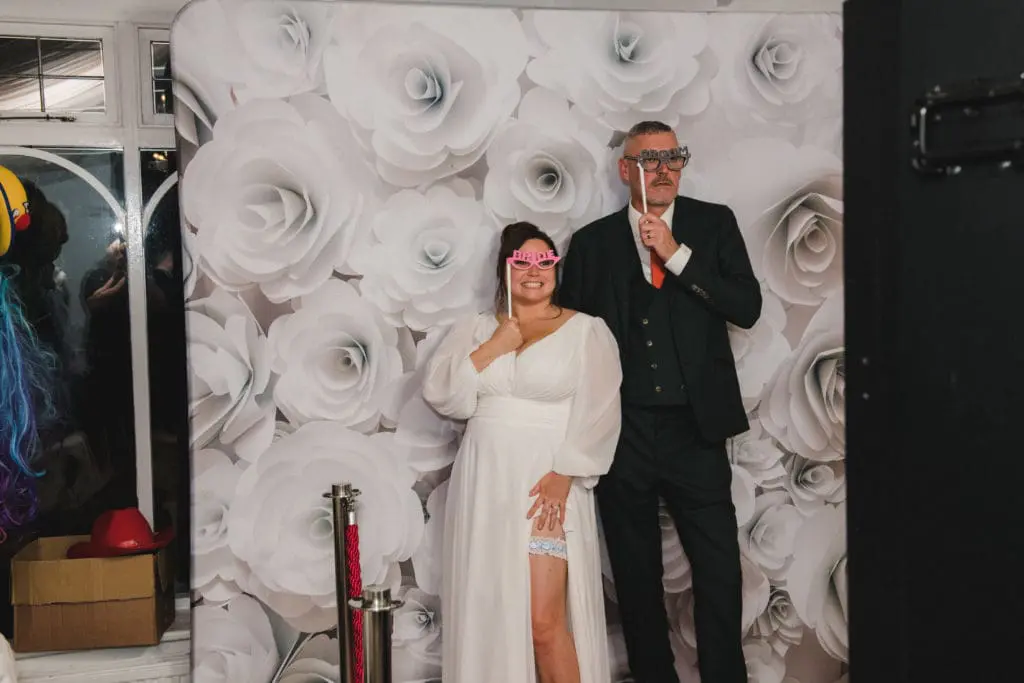 wedding photographer in Bedfordshire - Bride and groom standing in front of a white floral background holding bride and groom glasses to their face