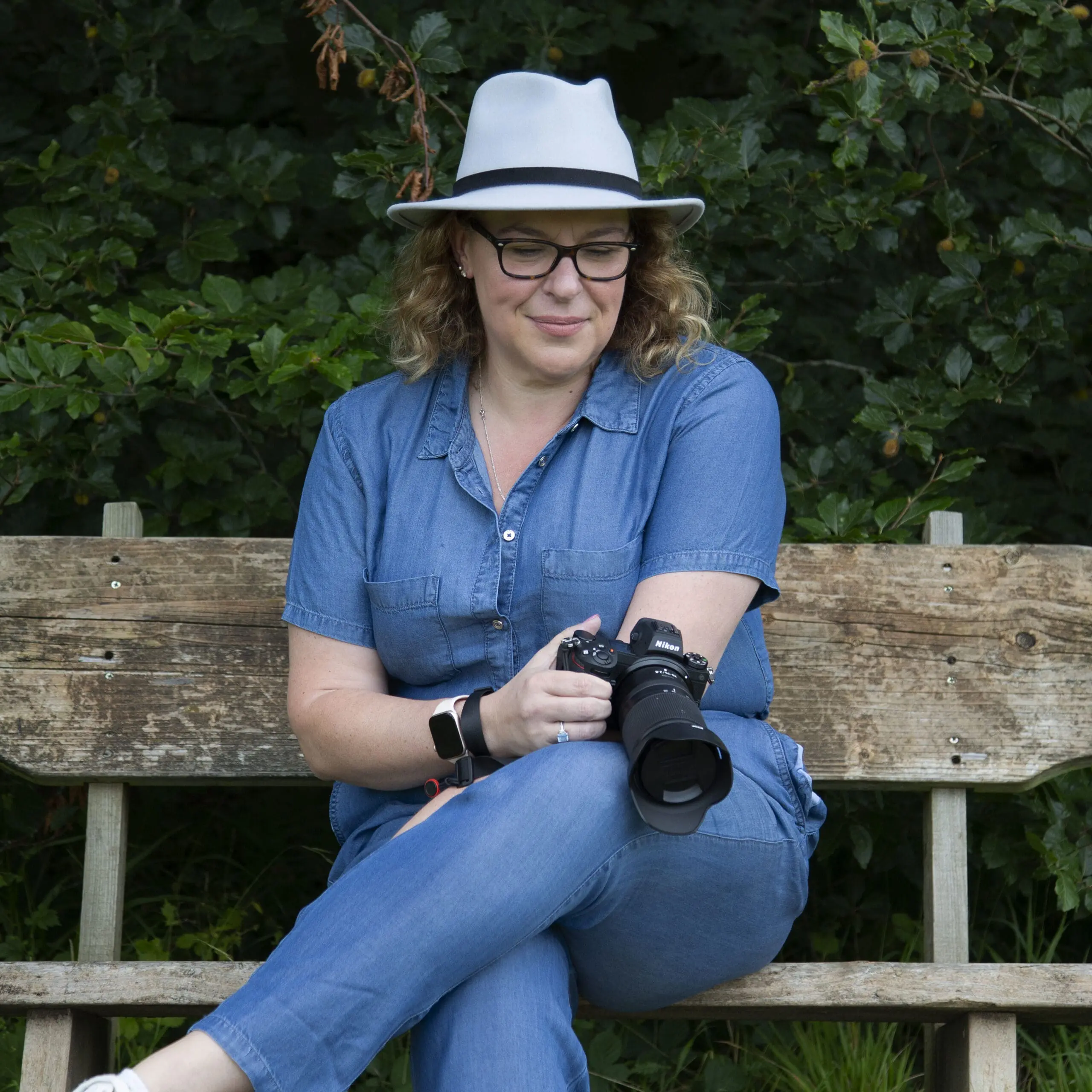 Catherine-22 Catherine Brooks Photography - Catherine a lady in a denim jumpsuit, sat on a wooden bench with her camera. She is wearing a grey fedora hat and black glasses