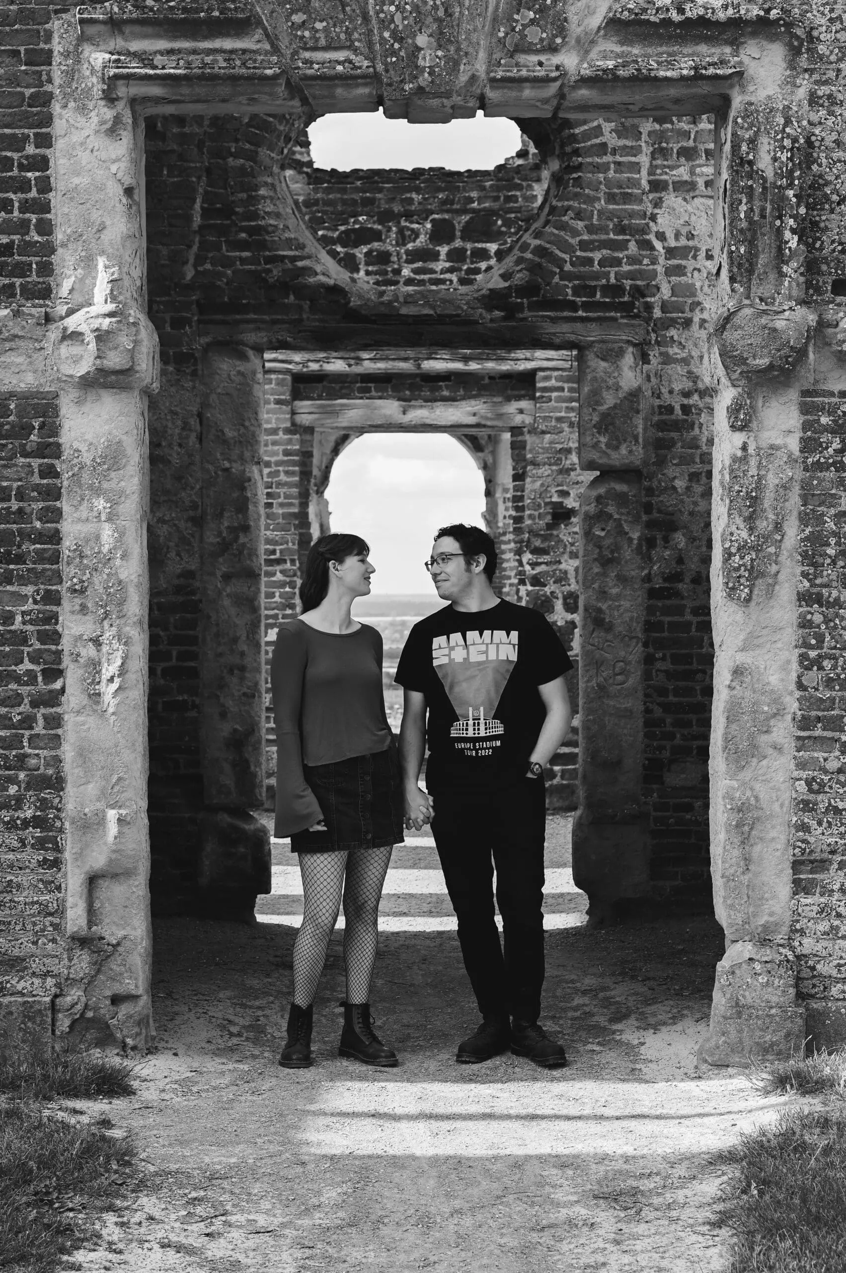 Houghton House Bedfordshire Engagement photography Engagement Photography Prices - a black and white image of a couple walking hand in hand out of a ruined archway