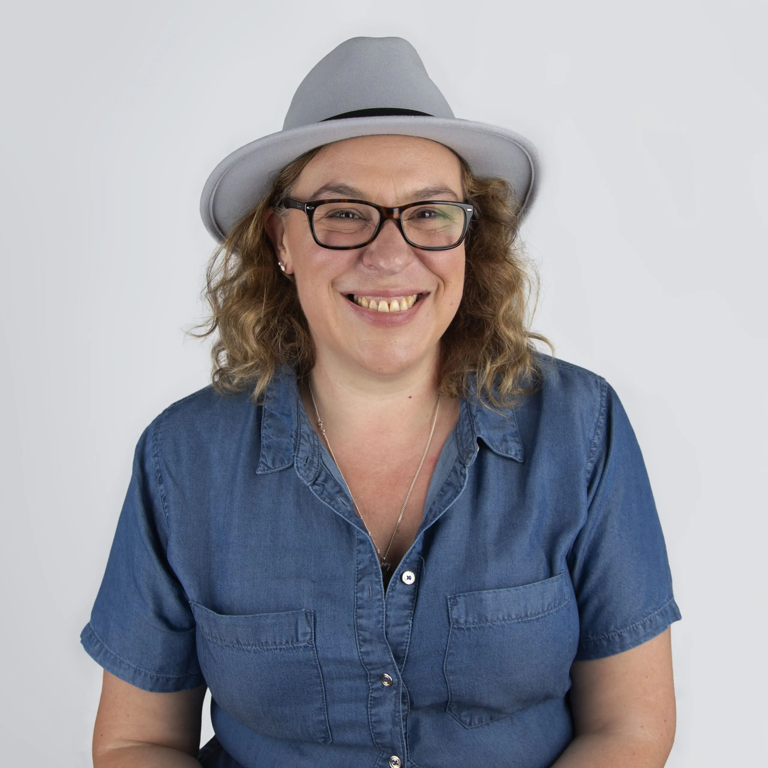 Bedfordshire Wedding Photographer Catherine Brooks Photography - Image of Catherine a lady wearing a denim shirt with curly mouse brown hair to the shoulder and glasses. She has a grey fedora hat on her hat and black glasses and is smiling