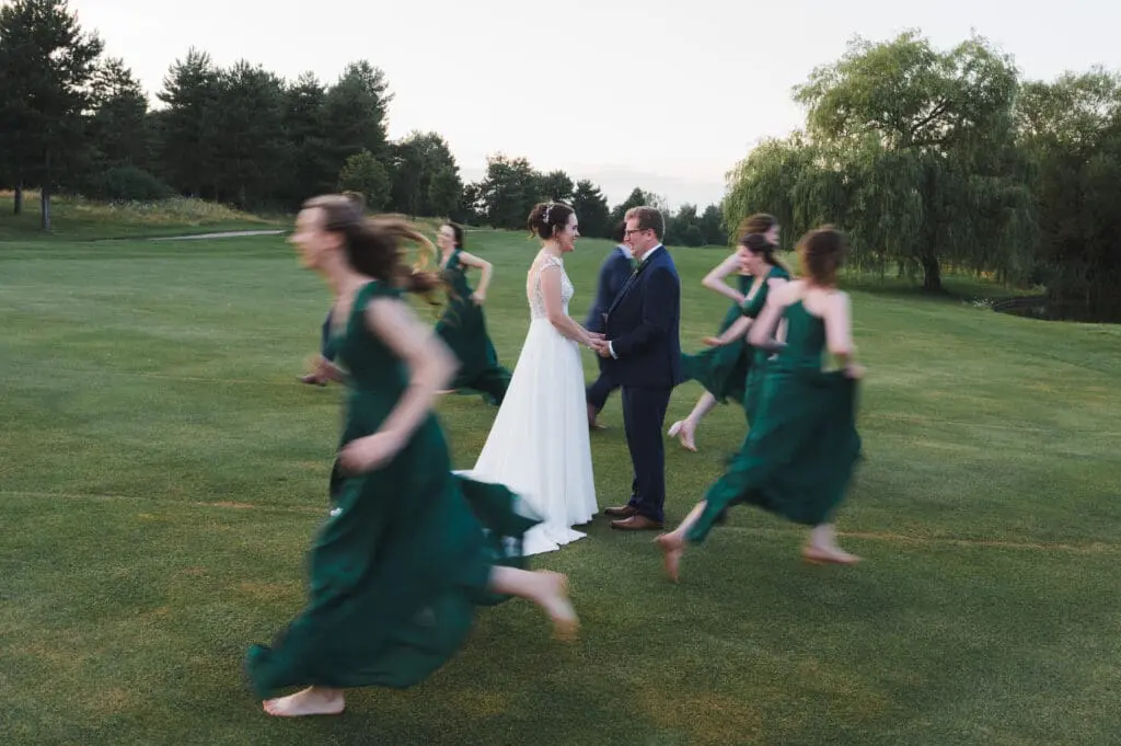 A bride and groom stand central in a field as their bridesmaids run past them  blurred
