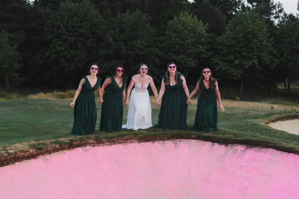 UK wedding photography trends include adding vibrant colours like this pink golf bunker, with the bride and bridesmaids getting ready to jump into it