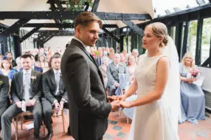 South Farm Wedding ceremony in a beamed room - the bride and groom are holding hands as guests look on