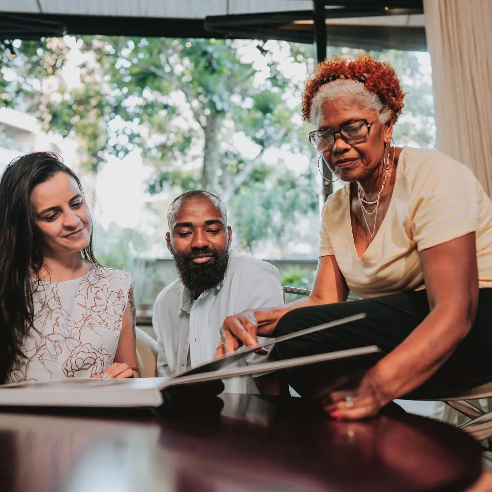 A dark-haired lady and a black man with a beard are looking through their wedding album keepsake with the groom's black mother in their lounge