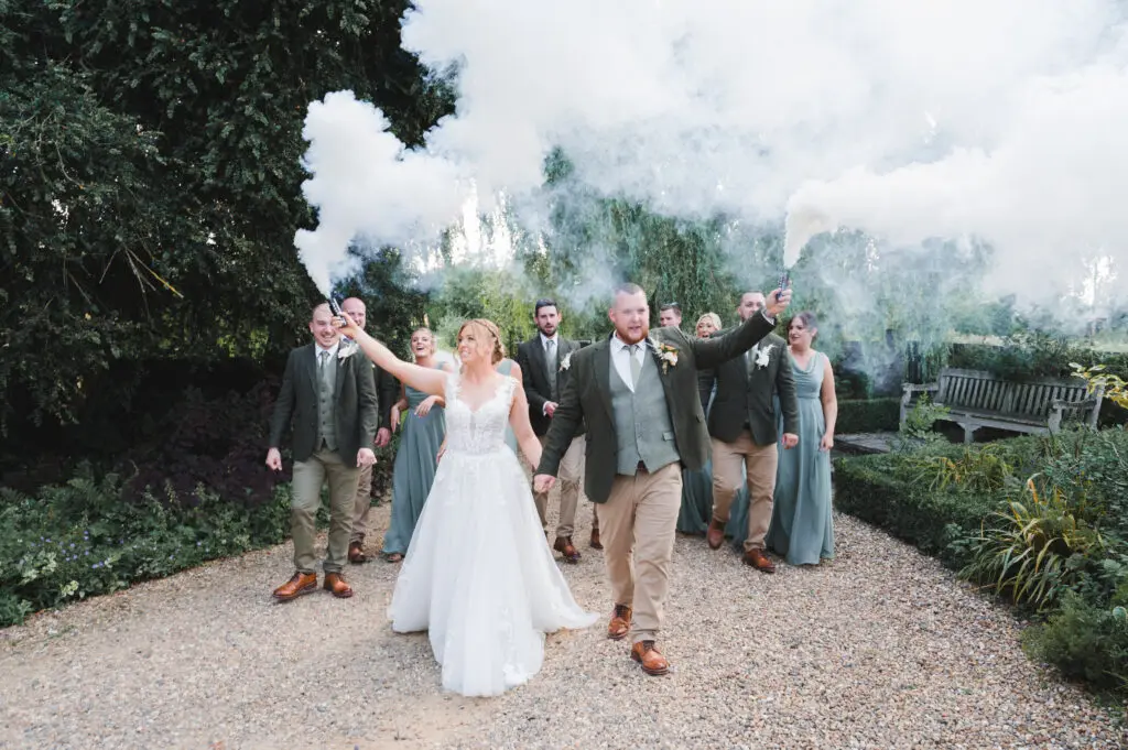 A bride and groom lead their wedding party along a pretty gravel path in the grounds of a house, holding white flares that release smoke