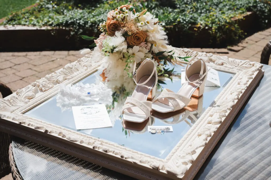 Wedding photography timeline - the bridal bouquet, shoes, garter, invite and jewellery placed on a mirror and photographed outside.