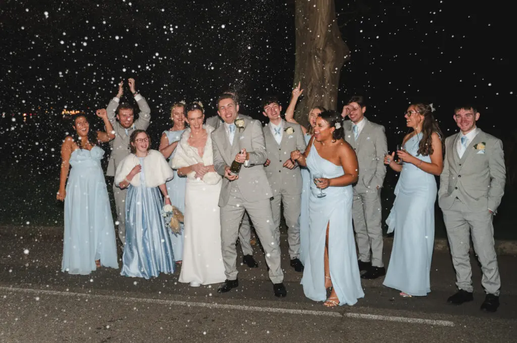 Look natural in your wedding photos like this wedding party of groomsmen dressed in grey 3-piece suits, bride and bridesmaids in pale blue getting covered in a champagne spray