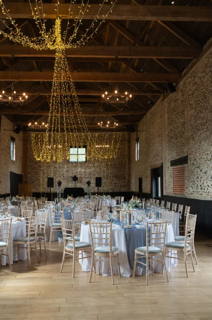 A barn wedding venue dressed in fairy lights with sky blue runners on the round white tables - a consideration when you manage a wedding budget