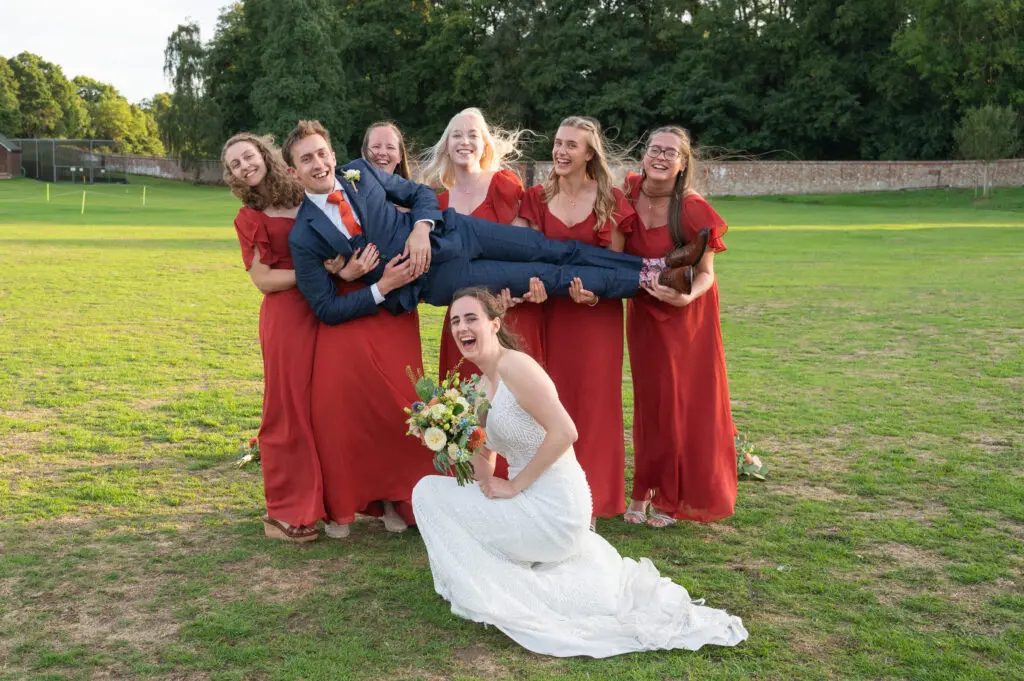 Eversholt Hall Weddings - 5 bridesmaids in dark burnt orange dresses are holding the groom in their arms, his bride is crouched beneath them laughing