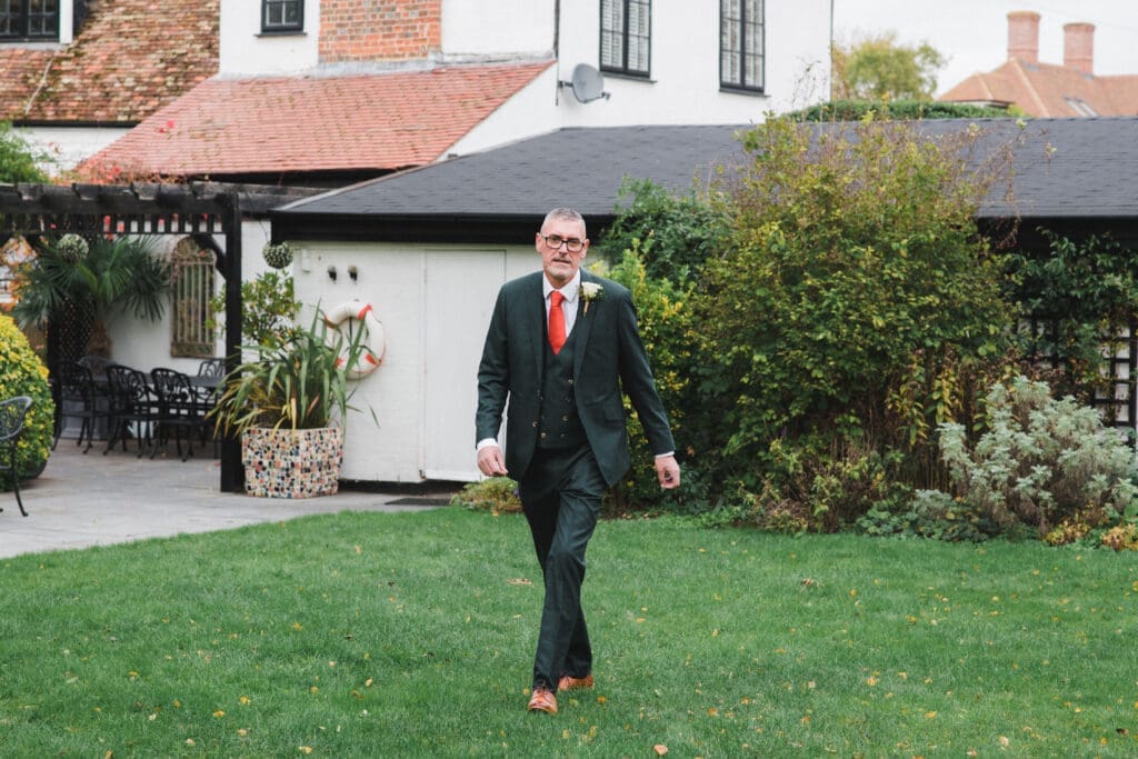 Sheene Mill Weddings in Cambridgeshire - Groom wearing an rust coloured tie is walking in the gardens with a white building behind him