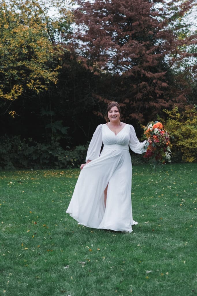 Sheene Mill Weddings in Cambridgeshire - A bride is walking towards the camera holding her dress in one hand and an autumnal bouquet in the other, there are trees in the background