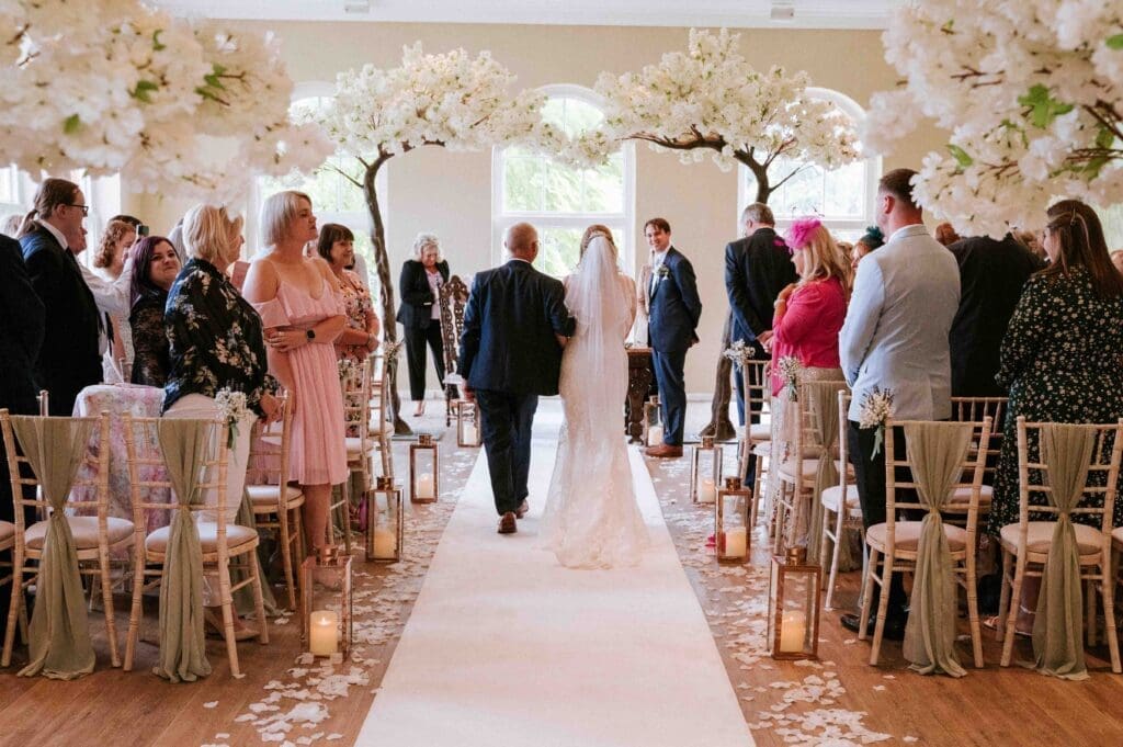 Braxted Park Wedding Photographer - The Orangery filled with white blossom trees. A bride walks down the aisle to meet her husband to be in front of their guests