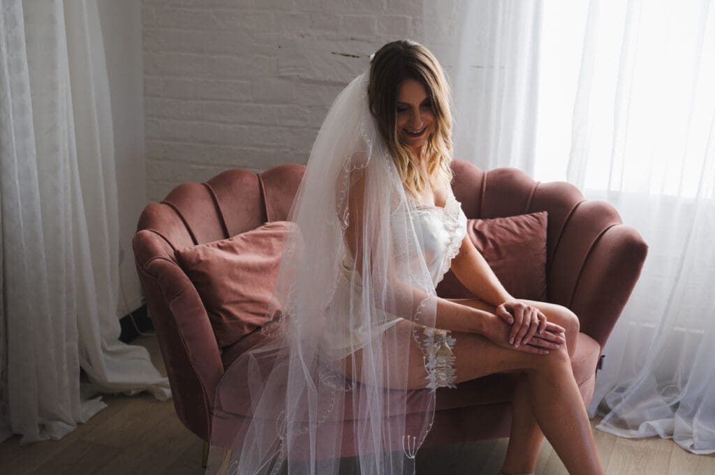 top boudoir poses for first-timers - a women in her bridal underwear and veil sits side on to the camera on a large pink chair looking down