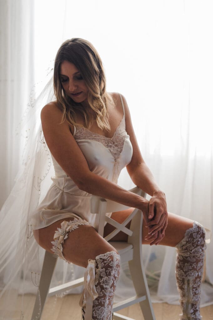 Bridal Boudoir gift - a bride in a white chemise and knickers, knee-high lace stockings, a garter and a veil, sitting astride a white chair
