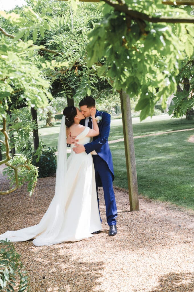 Seasonal Wedding Photography - summer wedding where the bride and groom are kissing underneath an arbour walkway covered in green foliage