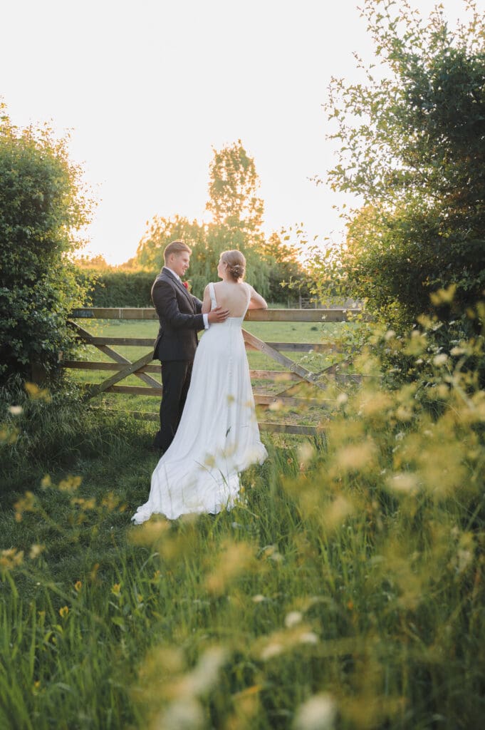 South Farm Wedding during Golden hour, the bride and groom look at each other, leaning on a field gate as the sun starts to set