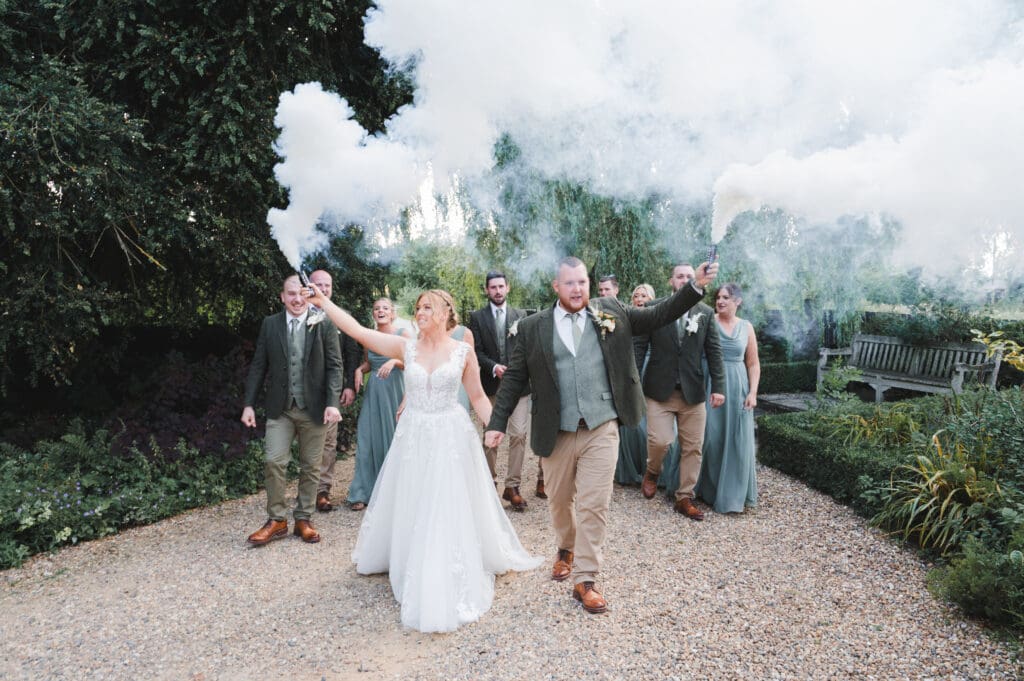 A bride and groom lead their wedding party along a pretty gravel path in the grounds of a house, holding white flares that release smoke
